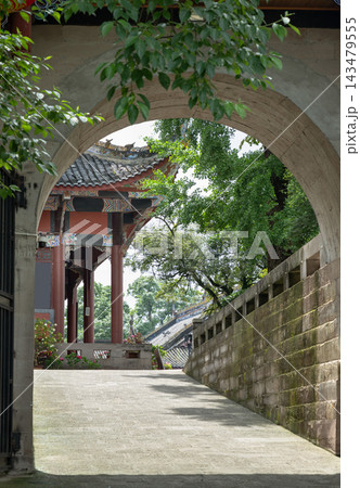 A view framed by a grand stone moon gate archway reveals a serene, sunlit path at the historic Dazu Rock Carvings complex in Chongqing, China. The foreground arch is delicately draped with vibrant A view framed by a grand stone moon gate archway reveals a serene, sunlit path at the historic Dazu Rock Carvings complex in Chongqing, China. The foreground arch is delicately draped with vibrant 143479555