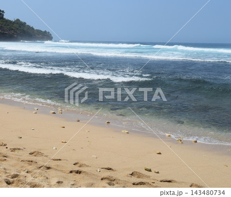 Scenic tropical beach with clear blue ocean water, gentle rolling waves, sandy shoreline, natural stones, and a lush green coastal headland under a clear blue sky. Scenic tropical beach with clear blue ocean water, gentle rolling waves, sandy shoreline, natural stones, and a lush green coastal headland under a clear blue sky. 143480734