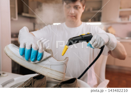 Man in protective gloves using handheld steam cleaner for deep cleaning and restoration of dirty white leather sneakers. Household chore and footwear maintenance concept. 143482210