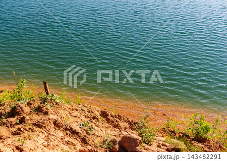 Serene turquoise lake water ripples gently against a rugged sandy shoreline featuring sparse green vegetation on a bright sunny day, creating a peaceful and natural coastal texture 143482291