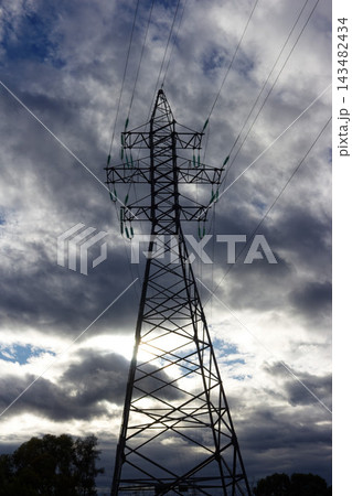 High voltage transmission tower silhouette standing against a dramatic cloudy sky with power lines stretching upwards towards the moody sunlight providing industrial energy distribution concept High voltage transmission tower silhouette standing against a dramatic cloudy sky with power lines stretching upwards towards the moody sunlight providing industrial energy distribution concept 143482434