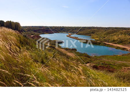 Panoramic view of a reclaimed sandy quarry featuring a calm turquoise lake nestled within grassy rolling hills and wild tall grass under a clear blue summer sky Panoramic view of a reclaimed sandy quarry featuring a calm turquoise lake nestled within grassy rolling hills and wild tall grass under a clear blue summer sky 143482525