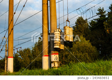 Concrete railway overhead line supports with tension weights hanging on steel cables against a backdrop of green forest under a soft sunset sky near train tracks 143483651