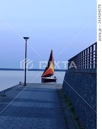 A sailboat with a bright sail approaches the waterfront at sunset. A sailboat with a bright sail approaches the waterfront at sunset. 143484079