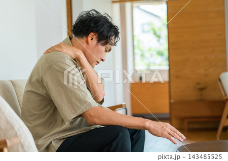 家のソファで肩を押さえて痛みに悩む40代男性。肩こりや首こり・筋肉疲労による体の不調のイメージ 家のソファで肩を押さえて痛みに悩む40代男性。肩こりや首こり・筋肉疲労による体の不調のイメージ 143485525