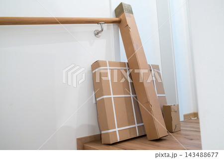 Three cardboard packages are stacked near a wooden handrail in a bright hallway. Delivery boxes of various sizes placed beside a staircase. Three cardboard packages are stacked near a wooden handrail in a bright hallway. Delivery boxes of various sizes placed beside a staircase. 143488677