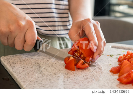 In a modern kitchen, a young woman is slicing fresh tomatoes with a knife on a cutting board. The meal preparation continues as she dicing the tomatoes for a healthy dish. In a modern kitchen, a young woman is slicing fresh tomatoes with a knife on a cutting board. The meal preparation continues as she dicing the tomatoes for a healthy dish. 143488695