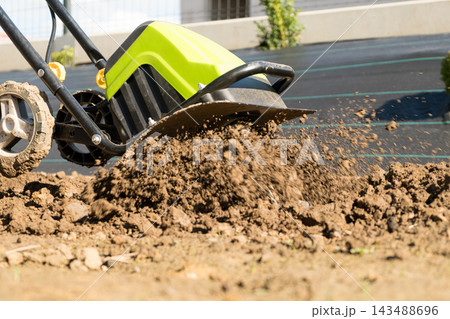 In a garden bed, a tiller is cultivating the soil to prepare it for crop planting. The tiller turns over earth, aiding in successful planting preparations. In a garden bed, a tiller is cultivating the soil to prepare it for crop planting. The tiller turns over earth, aiding in successful planting preparations. 143488696