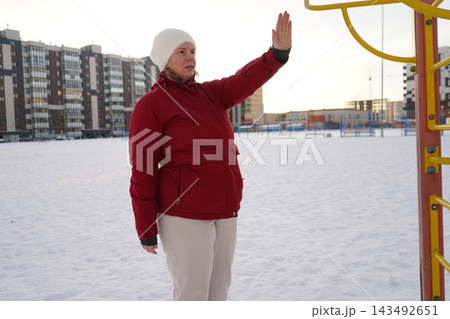 Side view of a mature woman in red jacket lifting arm to warm up at winter public sports ground. Side view of a mature woman in red jacket lifting arm to warm up at winter public sports ground. 143492651