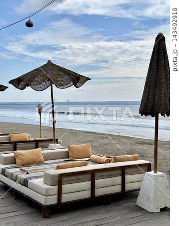 Cushioned beach daybeds face ocean waves beneath woven parasols in soft daylight. Cushioned beach daybeds face ocean waves beneath woven parasols in soft daylight. 143492918