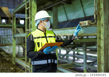 Worker in protective gear checking control panels and holding a clipboard at a waste recycling plant. Ensuring safety Worker in protective gear checking control panels and holding a clipboard at a waste recycling plant. Ensuring safety 143494345