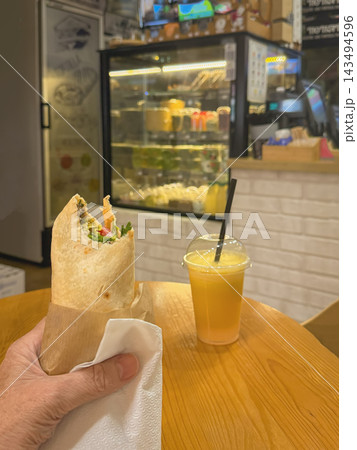 Hand Holding Tortilla Wrap Beside Orange Drink in Cafe. Hand holding a partially eaten tortilla wrap with lettuce and tomato beside Orange juice takeaway drink on wooden cafe table 143494596