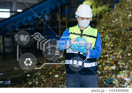 Asian engineer in hardhat using tablet with green energy and carbon neutral HUD graphics inside recycling factory 143494666