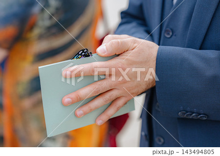 A man in a formal suit is holding a wallet indoors, possibly at an event or gathering. The image is a close-up shot focusing on the man's hands and wallet, highlighting the blue and white d. A man in a formal suit is holding a wallet indoors, possibly at an event or gathering. The image is a close-up shot focusing on the man's hands and wallet, highlighting the blue and white d. 143494805