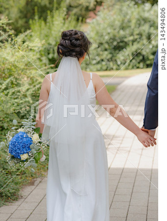 A bride walks hand-in-hand with her groom along an outdoor pathway, surrounded by lush greenery, captured from a third-person perspective behind the bride, creating a serene and romanticwed. A bride walks hand-in-hand with her groom along an outdoor pathway, surrounded by lush greenery, captured from a third-person perspective behind the bride, creating a serene and romanticwed. 143494806