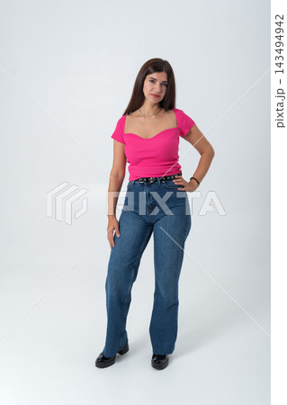Young woman standing confidently in a studio, wearing a bright pink ribbed top, blue high waisted jeans, and black shoes, presenting modern fashion and style 143494942