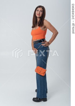 Young woman standing confidently in studio wearing orange crop top, blue jeans and matching orange shoulder bag, trendy casual portrait showcasing modern fashion and lifestyle style 143494943