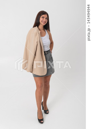 Smiling young businesswoman standing confidently in a white top and gray skirt, beige blazer slung over her shoulder, posing smart casual against a white studio background 143494944