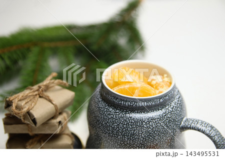 Horizontal close-up shot of a ceramic mug filled with mandarin slices, wrapped gifts, and a spruce branch. Christmas, winter holidays, and cozy atmosphere concept 143495531