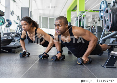 Black couple performing synchronized push-ups using dumbbells as handles on gym floor 143495747