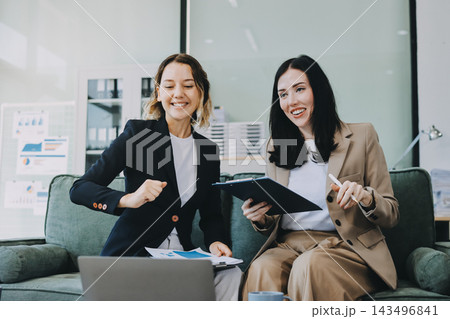Two smiling women are comfortably sitting on a couch, sharing a moment and collaborating on a tablet, working together in a home office environment 143496841