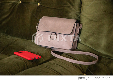 Crossbody bag and smartphone resting on a sofa. Everyday accessories for carrying essentials and staying connected. Casual fashion and personal technology. Modern lifestyle still life. Crossbody bag and smartphone resting on a sofa. Everyday accessories for carrying essentials and staying connected. Casual fashion and personal technology. Modern lifestyle still life. 143496887