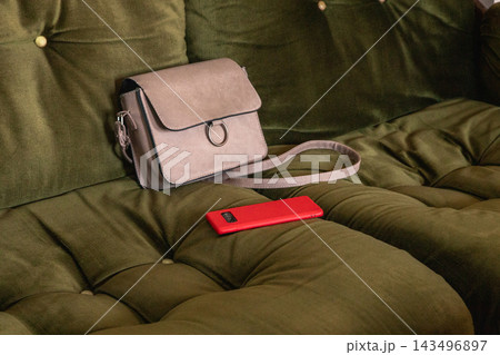 Crossbody bag and smartphone resting on a tufted sofa. Everyday accessories for mobile communication and personal style. Handbag and phone still life. Casual home lifestyle concept. 143496897