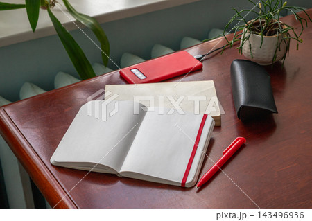 Open blank notebook, red pen and smartphone on polished wooden desk with houseplants. Home office workspace. Writing, planning and study concept. Stationery and productivity at work. 143496936