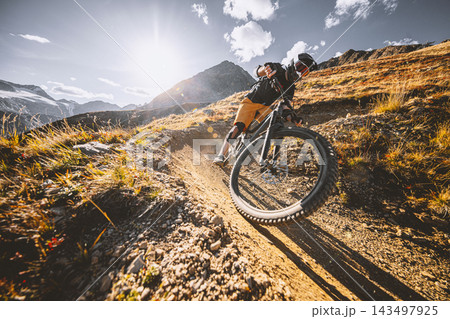 Mountain biker riding down a steep rocky alpine trail with dramatic mountains and a turquoise lake below, captured in warm sunlight during an adventurous high altitude descent ride. Mountain biker riding down a steep rocky alpine trail with dramatic mountains and a turquoise lake below, captured in warm sunlight during an adventurous high altitude descent ride. 143497925