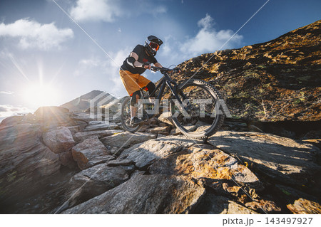 Mountain biker descending a rugged rocky alpine trail in bright sunlight, surrounded by dramatic mountain terrain and blue sky, capturing action, adventure, skill and extreme outdoor sport. Mountain biker descending a rugged rocky alpine trail in bright sunlight, surrounded by dramatic mountain terrain and blue sky, capturing action, adventure, skill and extreme outdoor sport. 143497927