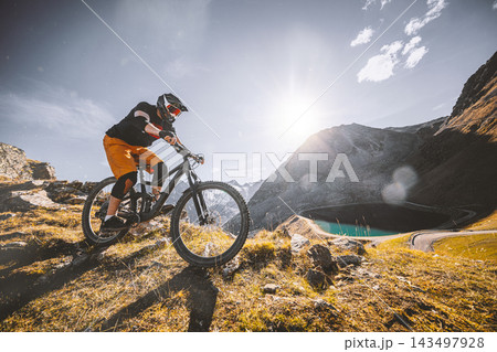 Mountain biker descending a rugged alpine trail above a mountain lake, surrounded by steep peaks and golden grass in warm sunlight, capturing adventure, speed and extreme outdoor sport. 143497928