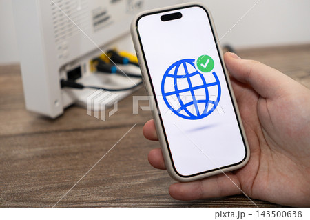 A hand holding a smartphone showing a global network icon with a checkmark, indicating active internet access beside a modern home router for wireless setup or tech troubleshooting A hand holding a smartphone showing a global network icon with a checkmark, indicating active internet access beside a modern home router for wireless setup or tech troubleshooting 143500638