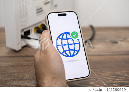 A hand holds a smartphone displaying a globe icon with a checkmark, symbolizing successful internet connectivity near a home Wi-Fi router to represent modern digital communication ease 143500639
