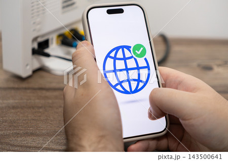 A person holds a smartphone displaying a globe icon with a checkmark while standing near a home internet router to confirm a stable network connection for digital communication tasks 143500641