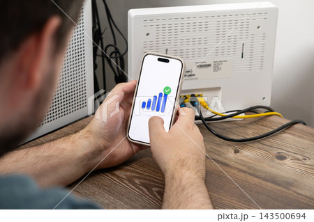 A man holds a smartphone displaying a full wireless signal icon while configuring a home router, illustrating successful internet setup and high speed connectivity in a modern home A man holds a smartphone displaying a full wireless signal icon while configuring a home router, illustrating successful internet setup and high speed connectivity in a modern home 143500694