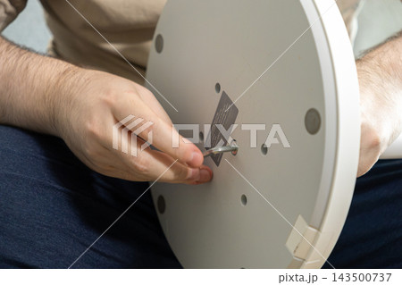 Person assembling a white pedestal fan base with an L-shaped hex key tool, demonstrating DIY home improvement skills and furniture construction in a bright residential indoor setting Person assembling a white pedestal fan base with an L-shaped hex key tool, demonstrating DIY home improvement skills and furniture construction in a bright residential indoor setting 143500737