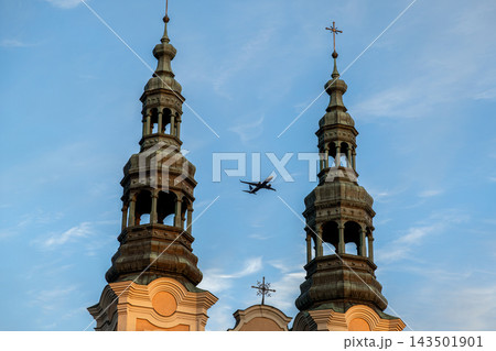 A passenger airplane flies between twin stone church towers under a clear blue sky, illustrating the juxtaposition of religious architecture and modern aviation travel in urban space 143501901