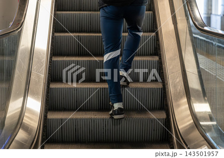 A person wearing casual blue athletic trousers walks up a metal escalator inside a modern building, representing daily urban commute, active lifestyle, and efficient transport flow 143501957
