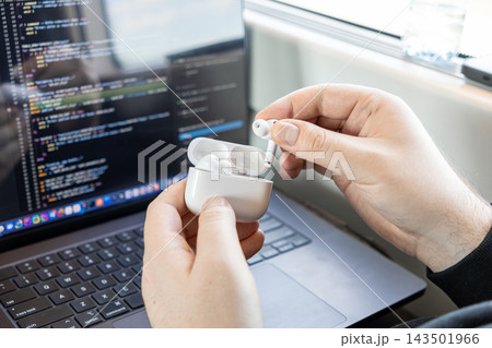 A developer taking wireless earbuds from a charging case while working on programming code on a laptop computer screen, representing modern remote software engineering and focus at work 143501966