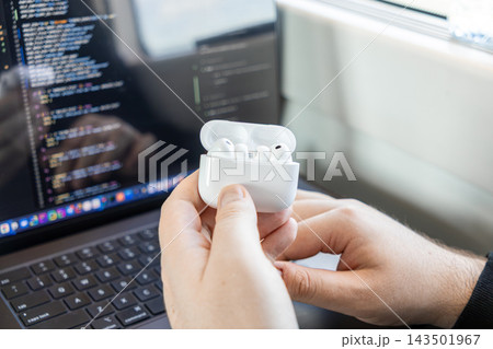 A programmer holding white wireless earbuds in a charging case against a laptop screen with programming code, symbolizing efficient modern remote tech development and digital lifestyle 143501967