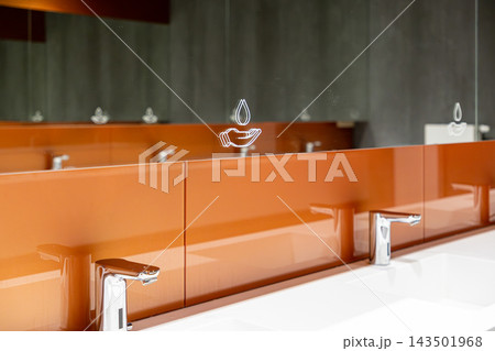 Modern public restroom vanity with orange paneling and mirror graphics, depicting clean water icons above automatic chrome faucets to promote hygiene standards in a professional facility 143501968