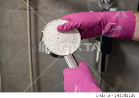 A hand wearing a pink rubber glove cleans a chrome shower head inside a tiled bathroom, representing home maintenance, hygiene routines, deep scrubbing, and professional cleaning A hand wearing a pink rubber glove cleans a chrome shower head inside a tiled bathroom, representing home maintenance, hygiene routines, deep scrubbing, and professional cleaning 143502139