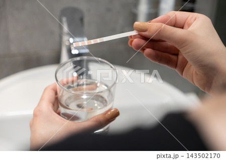 A woman testing water quality with a chemical paper strip, holding a glass against a bathroom faucet to ensure household health safety, purity, and standard tap maintenance assessment A woman testing water quality with a chemical paper strip, holding a glass against a bathroom faucet to ensure household health safety, purity, and standard tap maintenance assessment 143502170