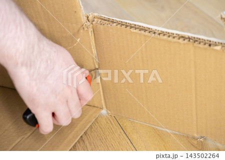A hand uses a utility knife to cut cardboard in a workshop, showcasing precise manual labor, home improvement, DIY crafting projects, and industrial packaging repair tasks at a desk A hand uses a utility knife to cut cardboard in a workshop, showcasing precise manual labor, home improvement, DIY crafting projects, and industrial packaging repair tasks at a desk 143502264
