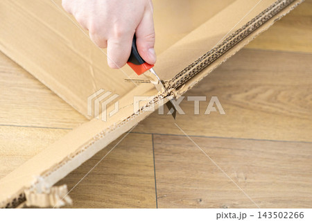 A hand uses a utility knife to cut through a thick brown cardboard sheet on a wooden floor, representing a DIY project, home renovation task, or package opening for shipping logistics A hand uses a utility knife to cut through a thick brown cardboard sheet on a wooden floor, representing a DIY project, home renovation task, or package opening for shipping logistics 143502266