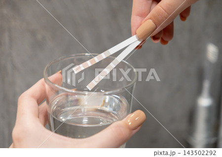 A hand holds a test strip over a glass of clear liquid to check water quality, representing a simple home health assessment procedure for safety analysis in a clean bathroom setting 143502292