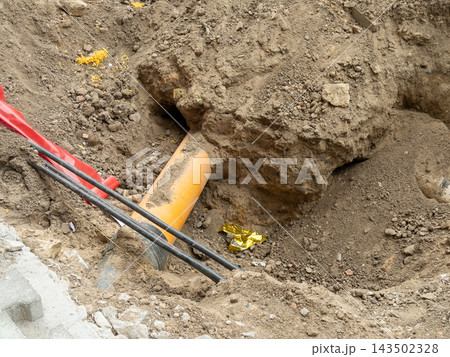 Orange sewer pipe and black cables exposed in a deep ground trench at a road construction site, representing utility infrastructure maintenance work during urban development project Orange sewer pipe and black cables exposed in a deep ground trench at a road construction site, representing utility infrastructure maintenance work during urban development project 143502328