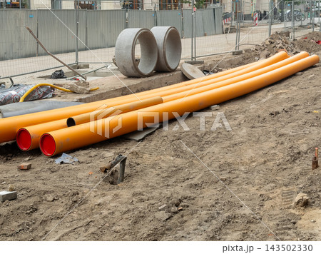 Bright orange plastic pipes for drainage rest on a muddy city construction site, highlighting infrastructure development, urban repair work, and modern utility installation projects Bright orange plastic pipes for drainage rest on a muddy city construction site, highlighting infrastructure development, urban repair work, and modern utility installation projects 143502330