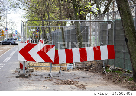 Red and white construction warning signs on a metal barricade near a city street sidewalk, signaling roadwork and safety caution to drivers and pedestrians in an urban environment Red and white construction warning signs on a metal barricade near a city street sidewalk, signaling roadwork and safety caution to drivers and pedestrians in an urban environment 143502405