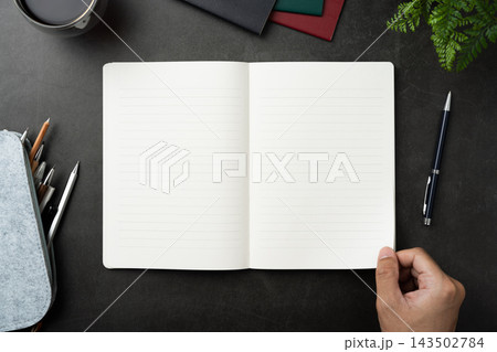 Top view of opened black hardcover notebook with hand hold page and office stationery on dark stone background with copy space 143502784